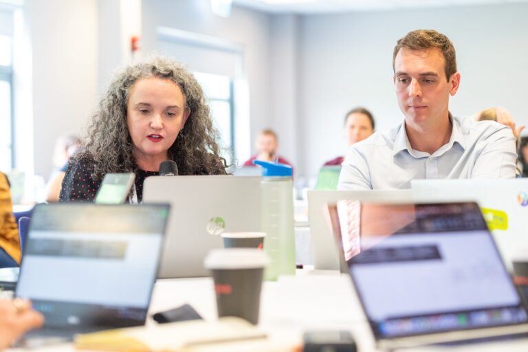 Focus on two people sitting around a crowded table where people are working on their laptops