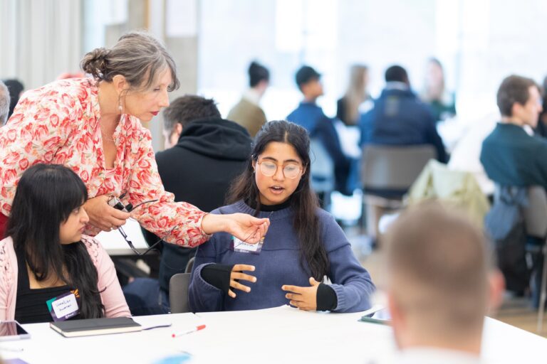 A workshop leader holds up a small microphone to a student participant who is giving feedback at a table of people