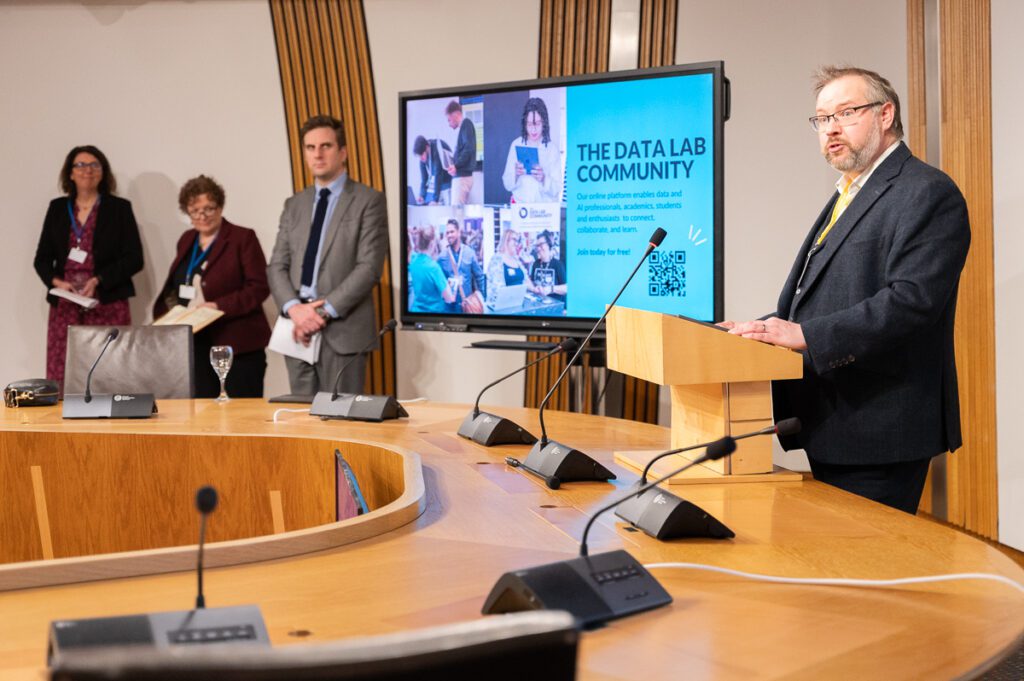 Brian Hills, CEO of The Data Lab, speaking at the lectern in the Burns Room in the Scottish Parliament, alongside Daniel Johnson MSP, Liz Whitefield from Hippo Digital and Dr Elizabeth Fairley from Talking Medicines.