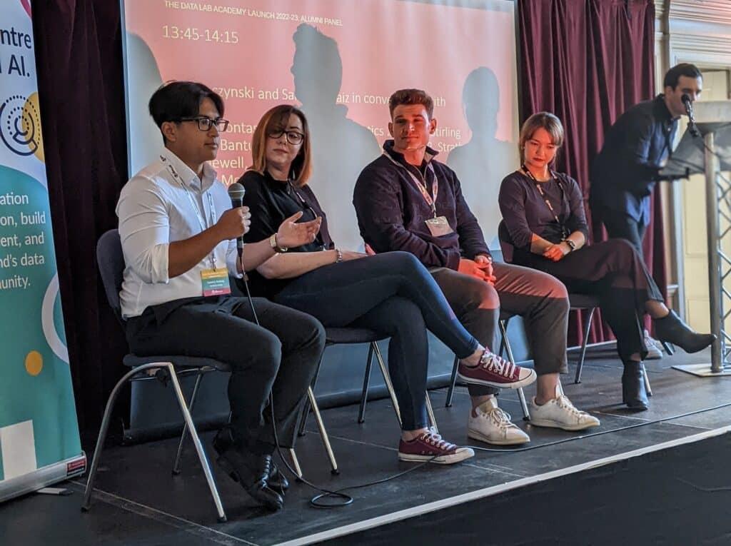 4 of the data lab academy alumni sitting on chairs in a row, giving a talk from a stage