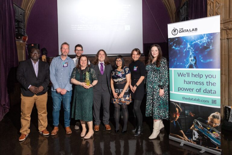 The Data Lab Academy at their celebraton event. A group of eight people stand in front of a Data Lab pull up banner all smiling.