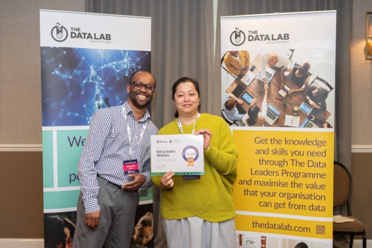 Dilraj Sokhi-Watson receives her graudation certificate at the Data Leadership graduation. She is wearing a green top, smiling brightly and standing in front of two promotional banners for the programme.