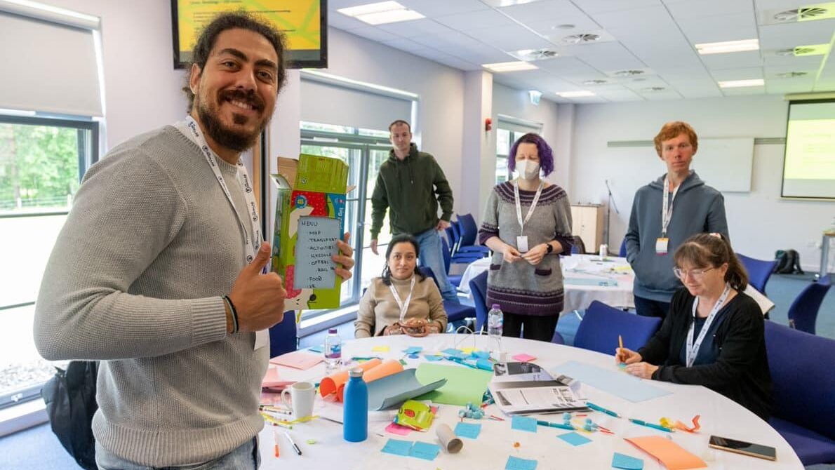 Six people working around a white round table that is covered in workshop material such as post-it notes, pens and paper. The man in the forefront of the image is posing giving a thumbs up. He has tanned skin, black curly hair that is tied up, dark facial hair and is wearing a light grey sweater and blue jeans.