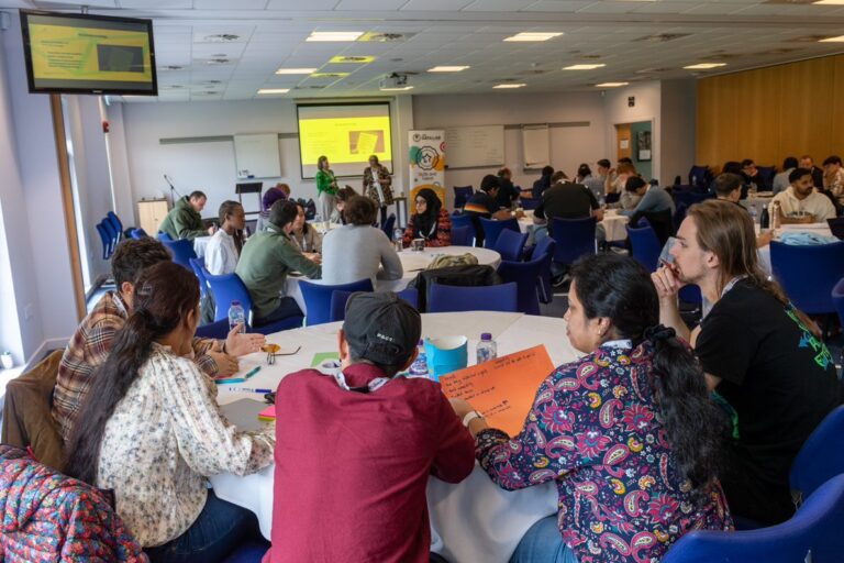 Students sitting at various tables in front of a presentation screen.