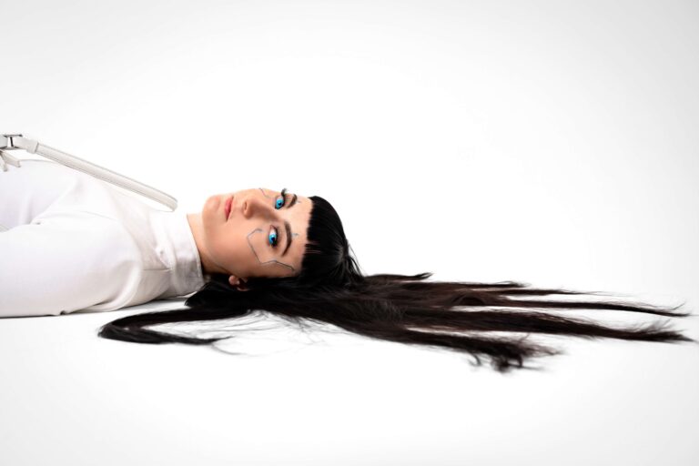 Kleo lying on her back on a white floor wearing ehite and looking sideways to the camera