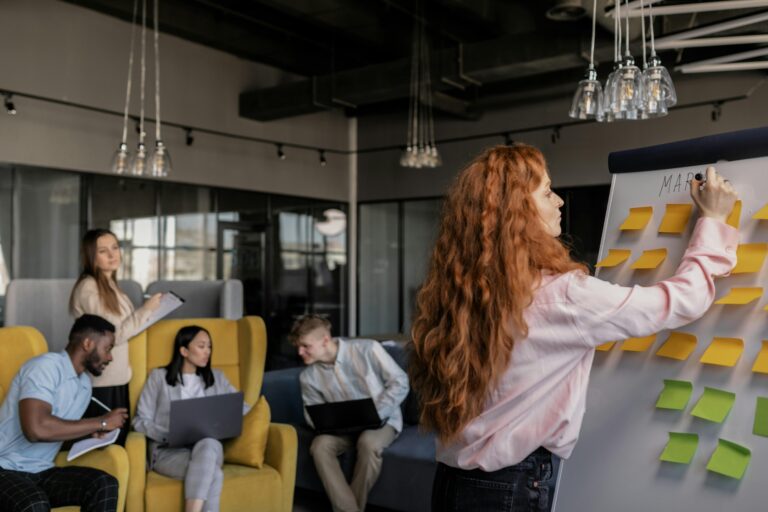 A diverse group of professionals in a modern office, brainstorming with sticky notes as a woman writes on a whiteboard.