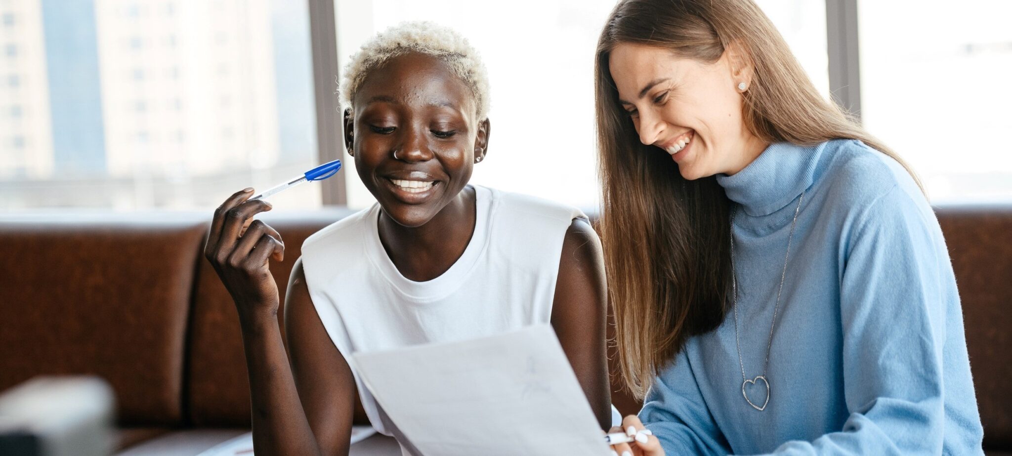 A young woman with radiant black skin and short, curly bleached blonde hair in a short-sleeved white top smiling and thinking with a blue pen resting on her cheek. She is looking down at a piece of paper held by a woman who looks in her 30's with long brown hair and white skin who is pointing at the paper with a big smile.