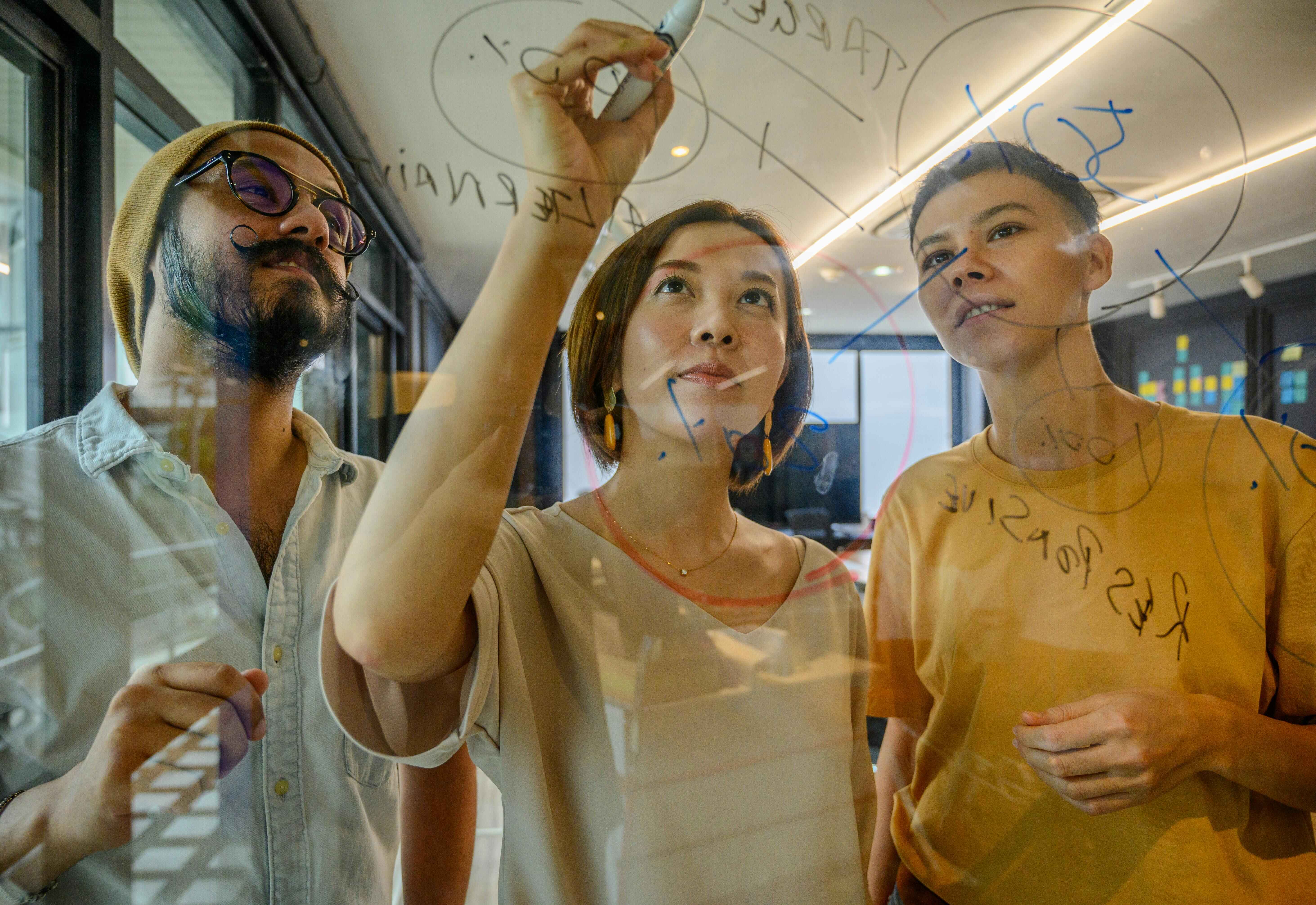 Three people working on a glass board actively trying to solve a problem.