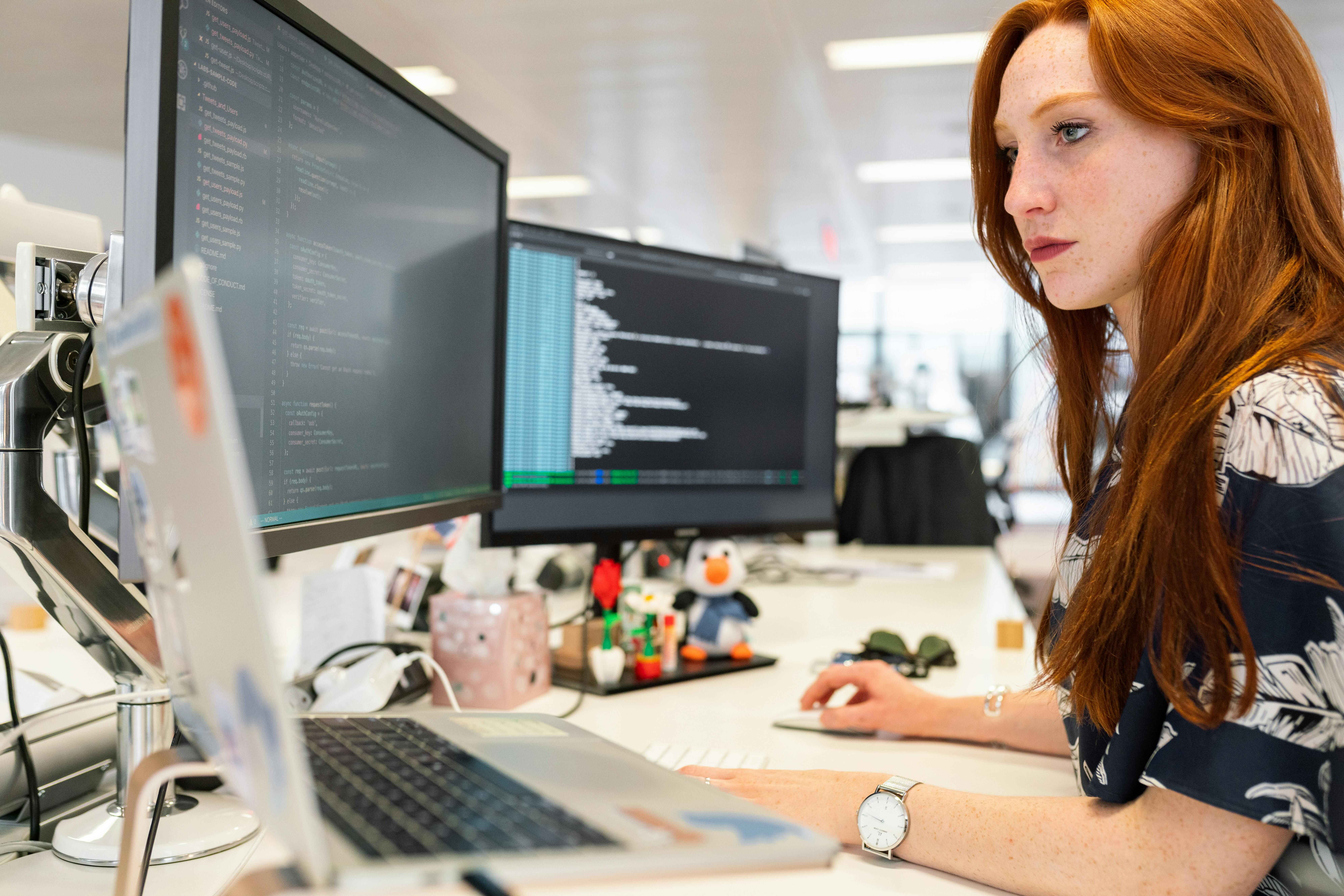 A woman with red hair sits at a desk with a laptop and two monitors - she is actively working with coding and data