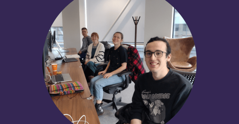 Four students sitting at a desk look up to the camera smiling