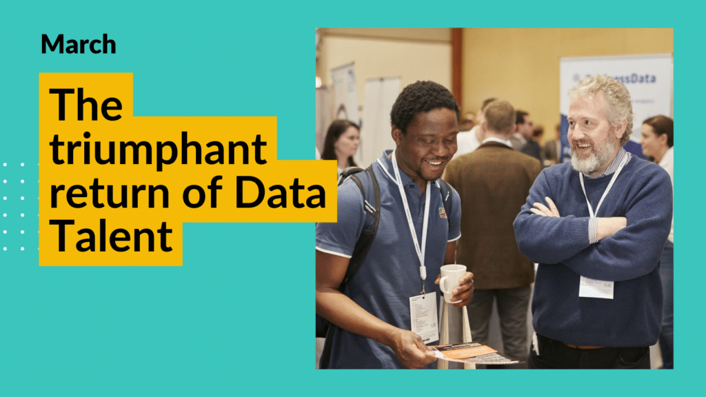 A student talks with an employer at Data Talent. They are smiling as they look down at a brochure. The man on the left is young with black skin and hair, he is wearing a navy shirt and a backpack. The man on the right has a light complexion and grey hair - he has his arms crossed and is smiling brightly. Overlay text reads "The triumphant return of Data Talent"
