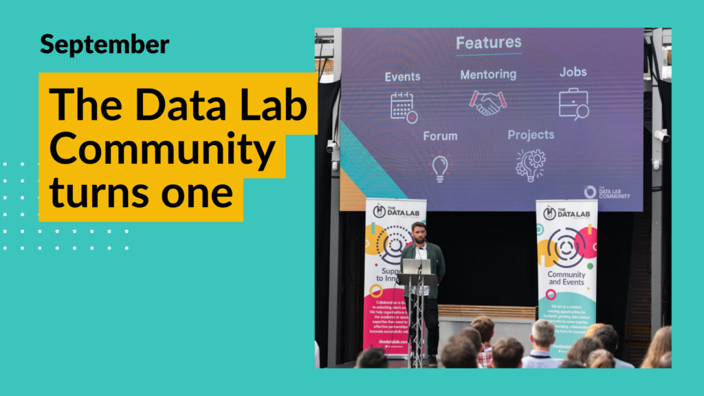 Image of Paul who runs The Data Lab Community standing in front of a large crowd. He is presenting on The Data Lab Community and standing at a podium with a presentation behind him. Overlay text reads "The Data Lab Community turns one"